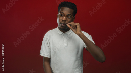 Tableau sur toile Young african american man wearing glasses and white polo with hand covering mouth for silence in red studio setting; silence