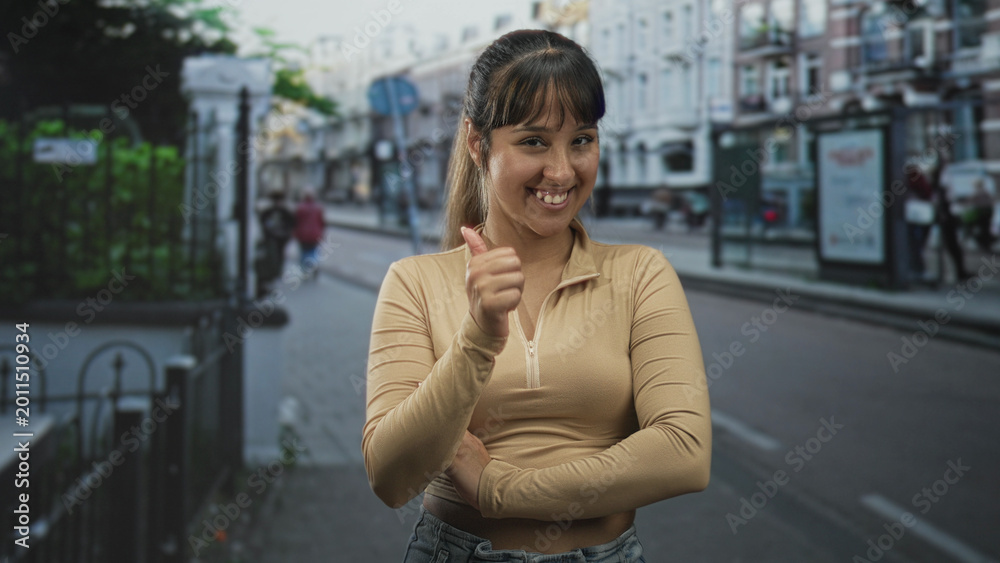 Fototapeta premium Woman with crossed arms and slight smirk wearing beige zip top on a city street near a bus shelter and railing; confidence assertiveness.