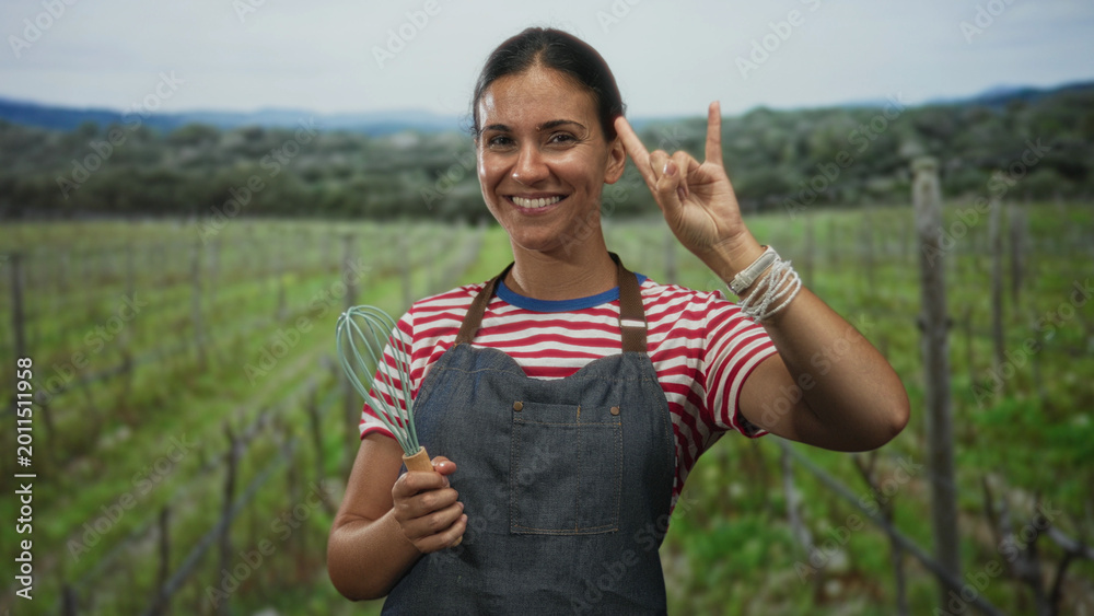 Fototapeta premium Woman wearing apron holding a whisk and making rock horns hand gesture in a vineyard field under open sky; rustic cooking joy.