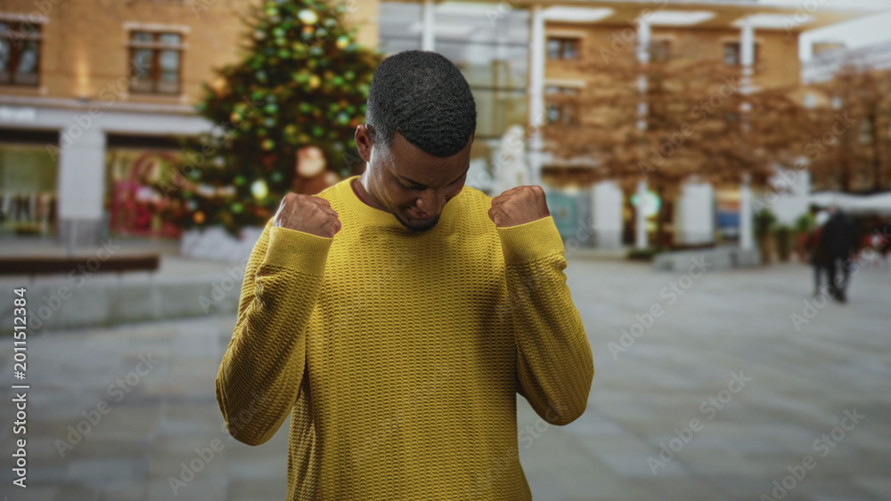 Obraz premium Young black man in yellow sweater raises his fists in a decorated street with a large christmas tree; quiet triumph.