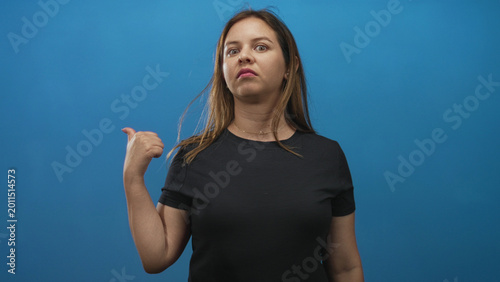 Foto Young caucasian woman in black t shirt points thumb over her shoulder while glancing sideways in a blue studio; annoyed