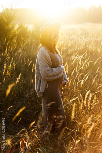 Stylish pregnant woman holding hands on belly and relaxing in sunlight in autumn park. Happy Mothers day! Beautiful mom hugging baby bump, and enjoying evening. Motherhood concept