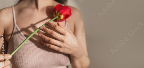 A woman holds a red tulip flower to her chest, against a light background. The concept of mammology, gynecology and women's health. Breast cancer banner copy space