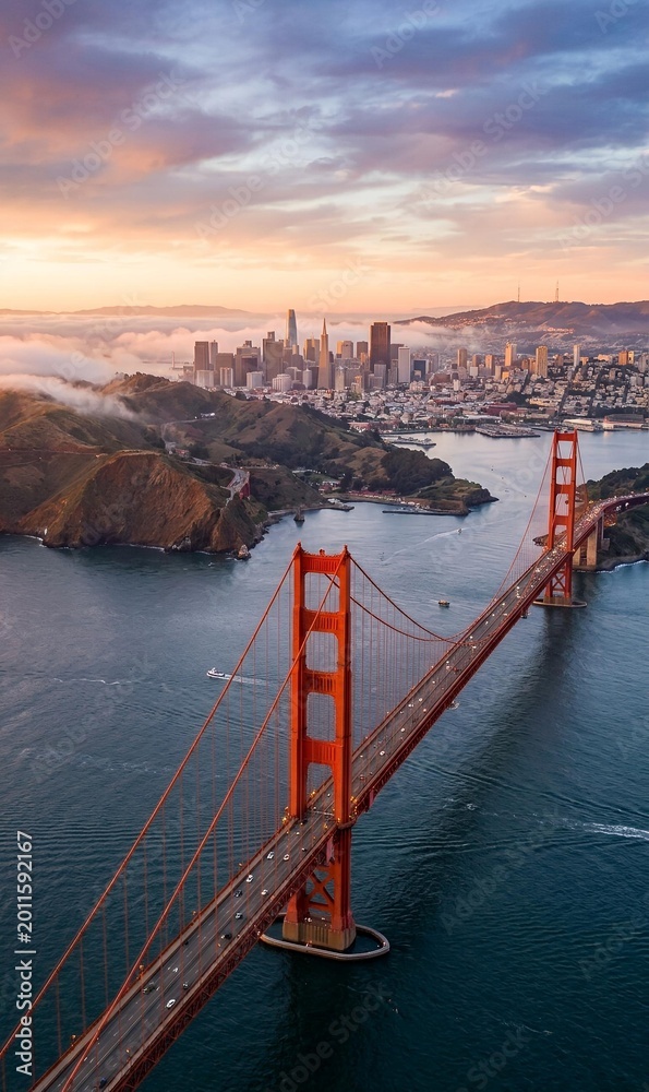 Obraz premium Aeriall vertical view of the Golden Gate Bridge and San Francisco skyline during a dramatic sunset with low fog.