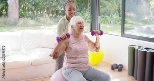 Female senior client in pink pressing weights African American trainer holding arms safely at home