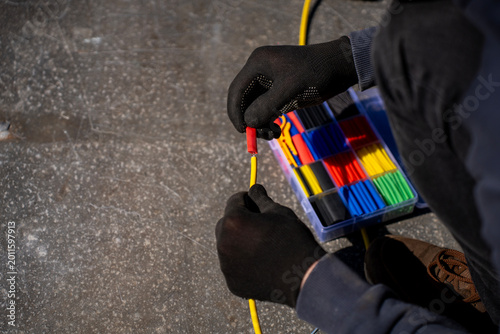 Obraz na plátně Gloved hands placing a red heat shrink tube over a yellow electrical cable beside a kit of colored shrink tubing outdoors