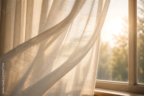 Sheer white curtain gently blowing in a soft breeze with warm morning sunlight streaming through a window and blurred trees outside.