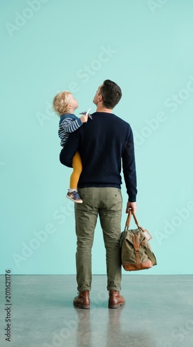 Back view of parent carrying toddler and duffel bag at airport isolated on light teal background