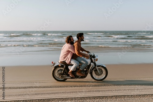 Two men riding motorcycle along sandy beach at shoreline