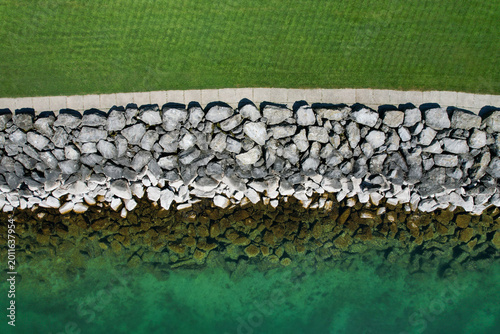 Aerial view of the rocky shoreline of Lake Geneva featuring a stone breakwater, a paved walkway, and lush green grass Morges, Vaud, Switzerland.