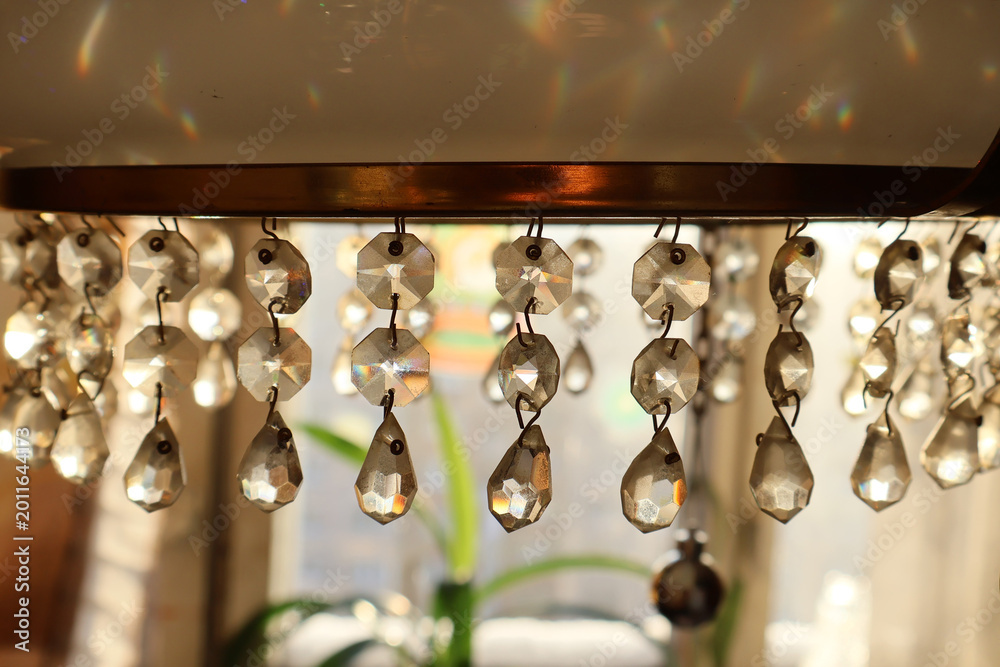 Fototapeta premium Close-up of hanging crystal prisms on a vintage chandelier, bathed in warm sunlight. The faceted drops sparkle, creating a mesmerizing play of light and rainbow reflections. The soft glow and intricat