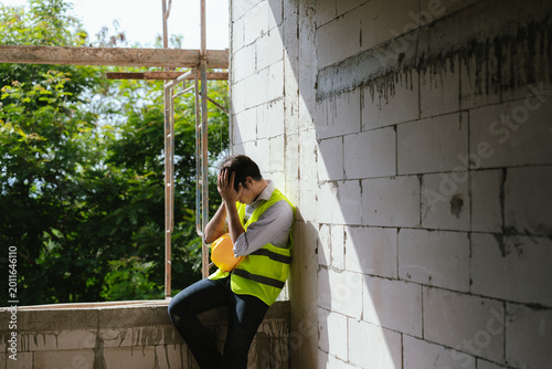 Valokuva Tired construction worker resting at a building site while holding a safety helmet, representing workplace stress, fatigue, and construction industry challenges