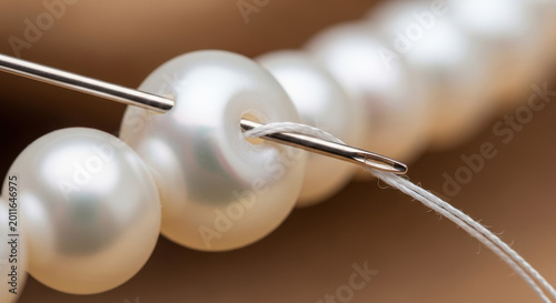 Necklace pearl being threaded with silver needle and white thread on brown background featuring pearls, jewelry, beads with threading and stringing
