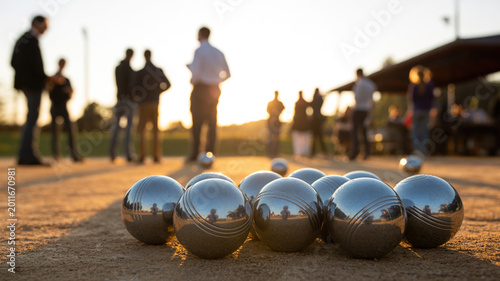 Metallic petanque balls arranged on sandy ground with blurred players in the background during sunset at an outdoor recreational area