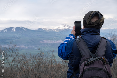 Obraz na płótnie Back view of traveler man with backpack on back admiring mountain landscape and taking photo using camera of mobile phone capturing snowy mountain peaks in distance