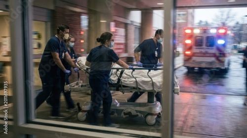 Medical professionals rush patient on gurney into hospital as ambulance arrives with flashing lights