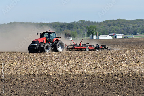 Red tractor  Pulling a  Double Fold Cultivator for spring field preparation with a farm scene in the background.