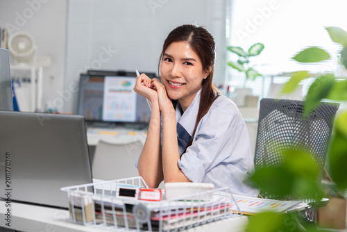 Happy cheerful young Asian woman smiling at camera sitting at bright modern office desk with laptop computer working professional portrait expressing feeling relaxed and content
