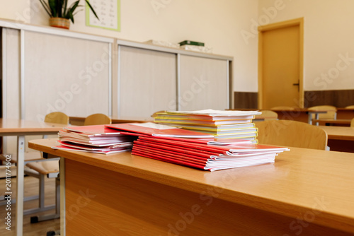 Stacks of notebooks on wooden desk in empty classroom with window background. School supplies on table in class. Back to school concept, elementary education, teacher workspace, learning, nobody