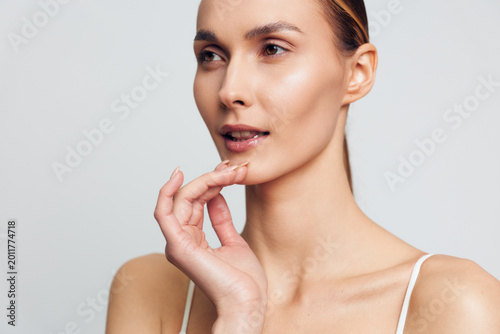 Young woman with clear skin gently touching her chin, soft natural light, minimal makeup, calm expression, smooth complexion, neutral background, skincare concept
