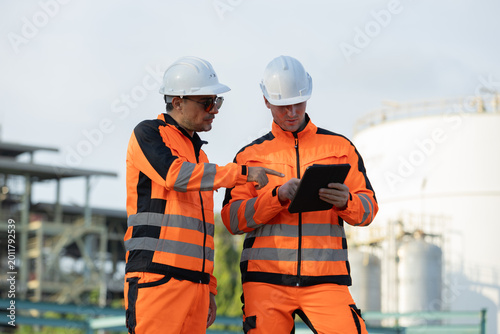 Two engineers in orange safety gear and helmets discuss work using a tablet at an industrial facility.