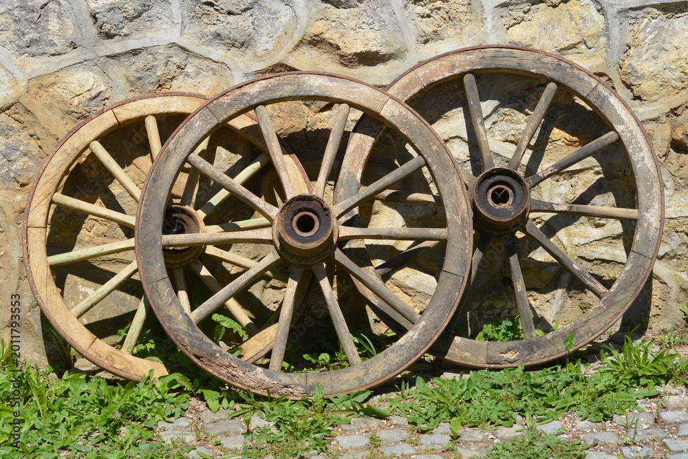 Obraz premium Three weathered wooden carriage wheels leaning against a rustic stone wall on a sunny day in a traditional courtyard