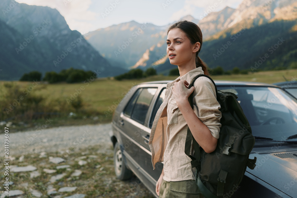 Naklejka premium Smiling woman with backpack beside a car in a valley with mountains in the background, wearing a casual jacket, enjoying an outdoors lifestyle, travel and adventure vibe under natural light.