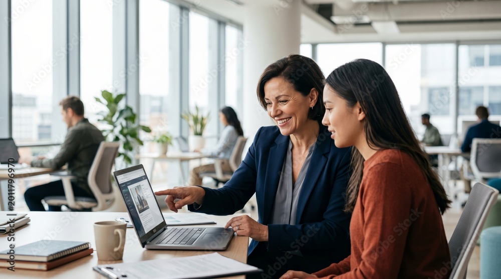 Fototapeta premium Two women reviewing business analytics on laptop in modern office. Teamwork, data analysis, and professional collaboration concept.