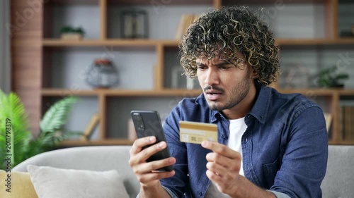 Frustrated young man discovered fraud entering credit card number on mobile phone sitting on sofa in living room at home. Upset shocked male became victim of deception, money had been stolen. Close up
