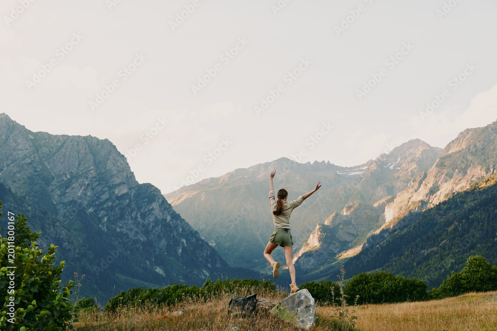 Obraz premium Young woman stands on a rocky outcrop with arms raised in celebration, framed by sunlit alpine valley and distant mountains, conveying freedom and a sense of adventure.