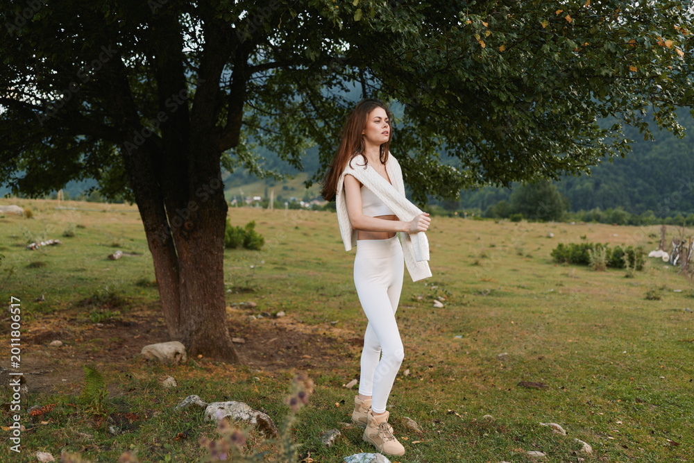 Fototapeta premium Young woman in white outfit standing near a tree in a green meadow. Outdoor lifestyle scene with nature and calm mood during daytime.