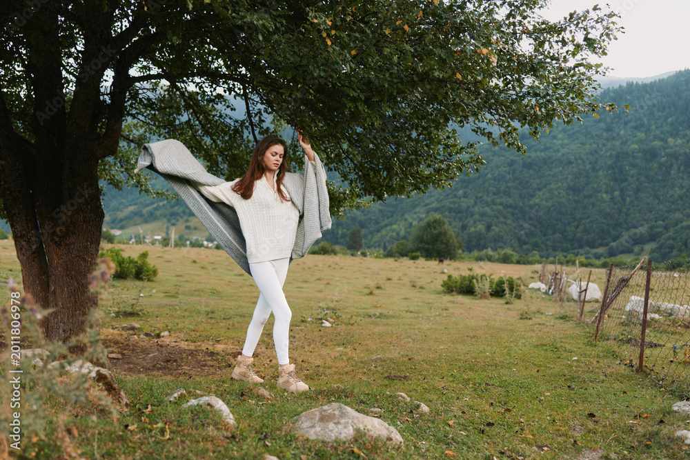 Fototapeta premium Young woman in white outfit posing outdoors under a leafy tree in a rural field. Lifestyle photography showing freedom, nature, and calm, with soft daylight and relaxed movement.
