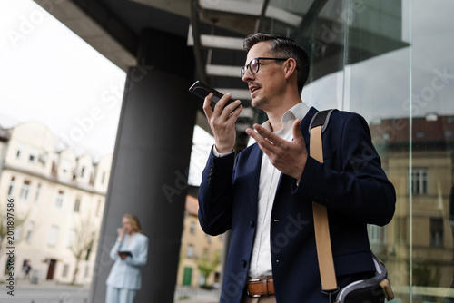 Man Using Voice Recorder Outside a Building While a Woman Checks Her Phone in the Background in the City on a Cloudy Day