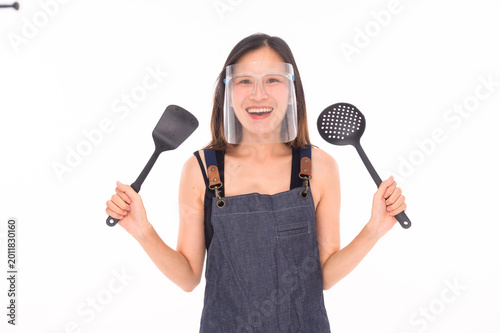 Valokuva A smiling woman wearing a face shield and apron holds two kitchen utensils, ready for cooking