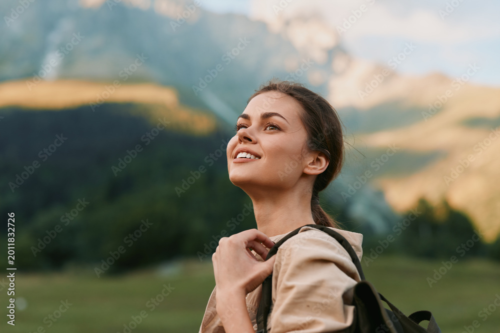 Naklejka premium A woman outdoors with a backpack smiles in a sunny landscape, embodying travel and wellness. A wide mountain meadow and blue sky form a hopeful setting for lifestyle exploration.