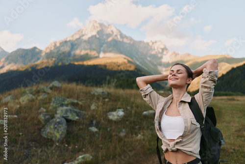 Young woman with a backpack stands in a mountain landscape, smiling with arms raised as sunlit peaks rise behind her and a wide blue sky. Under clear weather and fresh air.