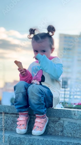 Cute little girl eating an ice cream during a city walk while sitting on outdoor steps with apartment buildings in the background. Happy childhood and summer leisure concept