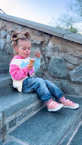 Little girl sitting on steps and eating ice cream during a walk in a residential city neighborhood. Summer leisure, childhood, and family lifestyle concept