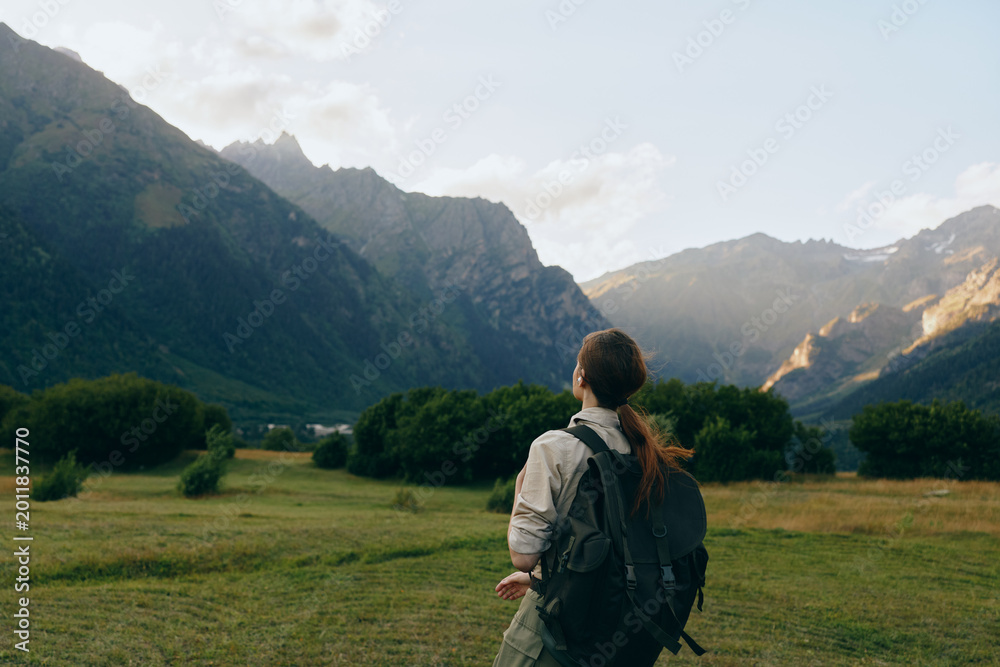 Naklejka premium Young woman with backpack stands in a sunlit valley, gazing toward distant mountains, capturing a moment of exploration and calm amid nature.