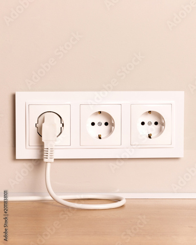 White power socket with electric plug inserted in wall outlet. Modern triple receptacle on light beige wall with cable curving on wooden surface