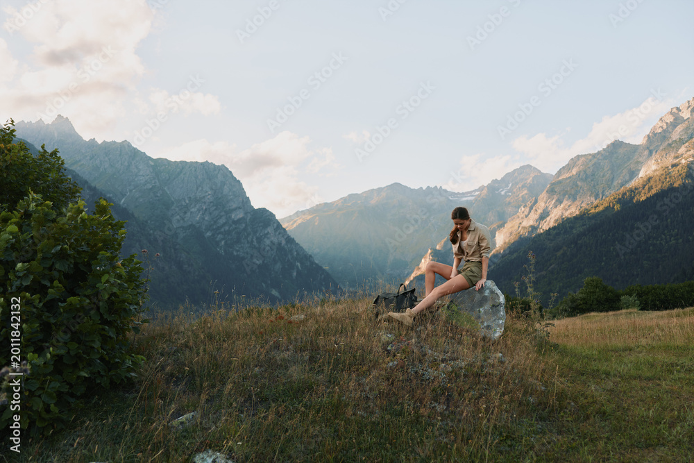 Naklejka premium Woman enjoying outdoor lifestyle in a mountain meadow, sitting on a rock and gazing at distant peaks, casual clothes, peaceful mood, warm sunlight, natural landscape and solitude inspires calm.