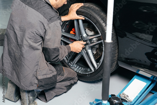 Принти на полотні Mechanic repairing car wheel in auto repair shop using lift jack and electric drill to loosen the bolts