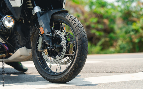 Close-up of a motorcycle front wheel with dual-sport tire and disc brake, parked on a street with greenery blurred in the background.