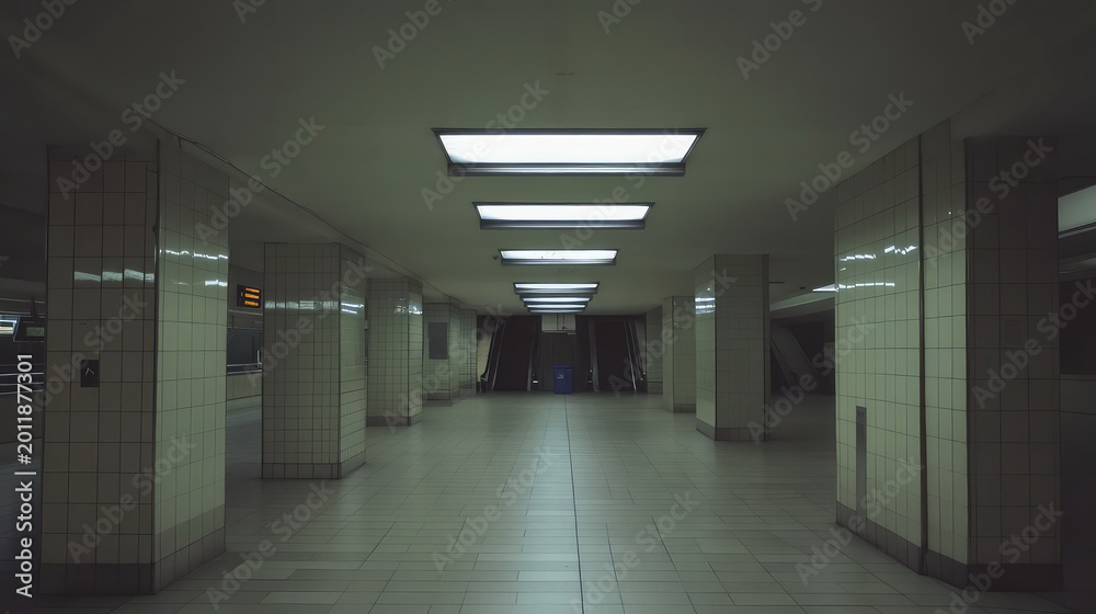 Fototapeta premium Empty underground metro platform with symmetrical tiled pillars, glowing rectangular ceiling lights and distant escalators, conveying urban transit, solitude and waiting