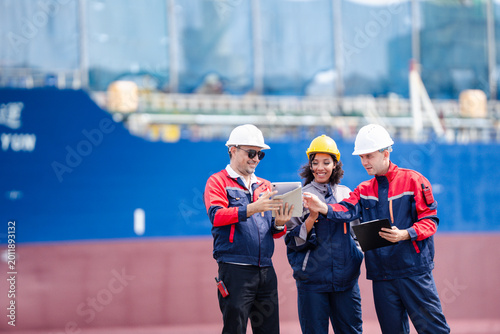 Industrial engineering team in safety helmets discussing operations and logistics management at shipping port with cargo vessel background and teamwork concept