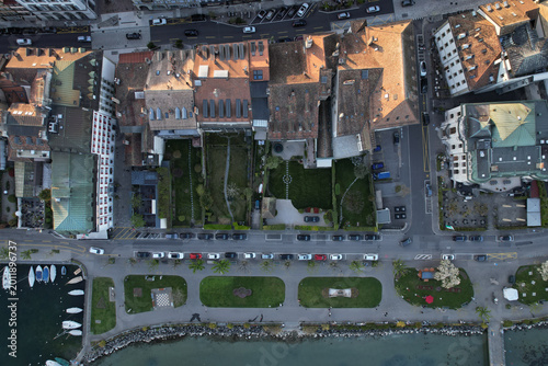 Aerial view of historic buildings with red-tiled roofs, lush private gardens, and people walking along the lakeside promenade Morges, Vaud, Switzerland.