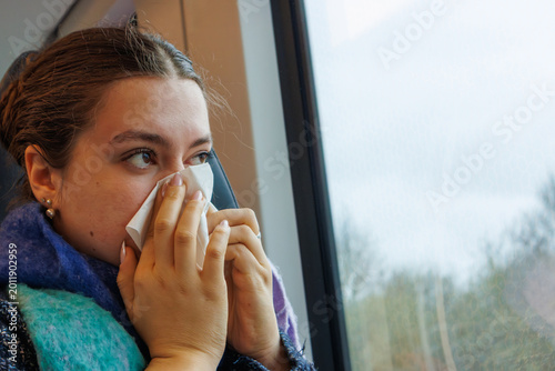 Close up of a teenage girl sitting by a train window and sneezing into a paper tissue. Copy space. Selective focus
