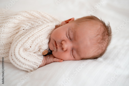 newborn baby sleeping on a white bed at home, close-up portrait of a newborn baby sleeping in a white knitted bodysuit, space for text