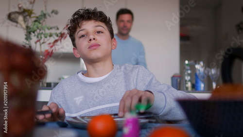 Boy sitting at dining table during family meal, looking ahead with content expression while food and fruit are placed on table in cozy home kitchen