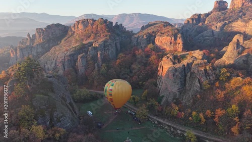 Wide aerial view of a hot air balloon on an open launch field surrounded by rocky cliffs and autumn trees.